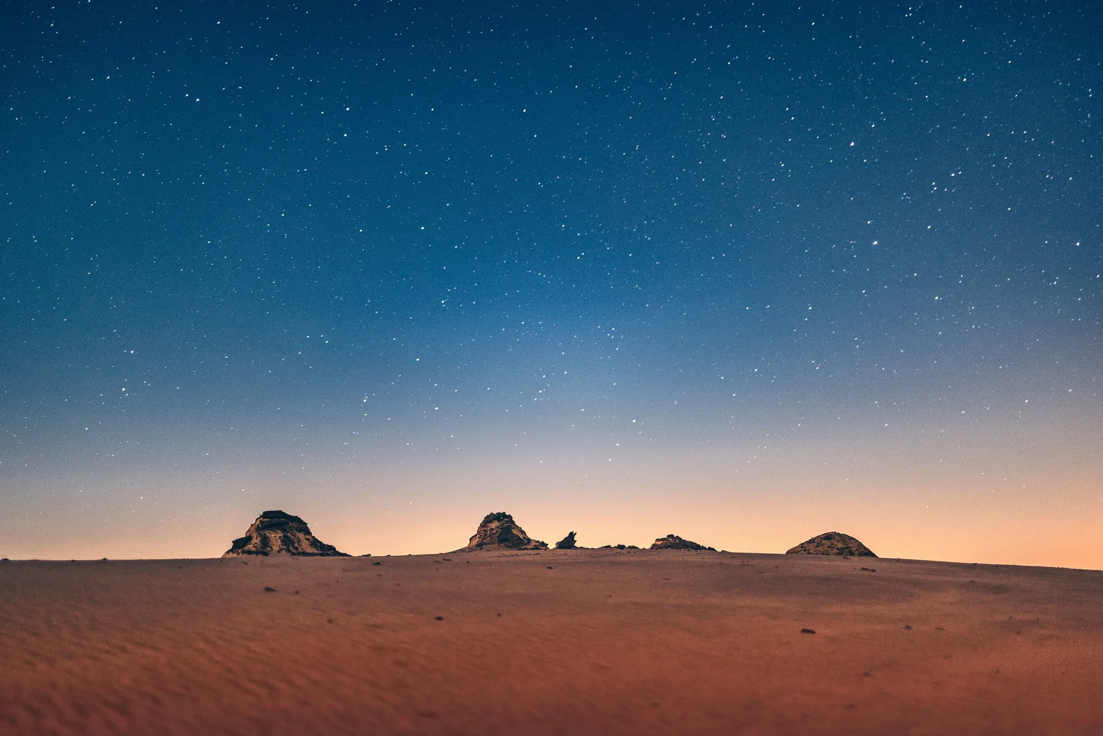 Sous un ciel étoilé, des dunes désertiques sombres et de petits affleurements rocheux (photographie représentative d'un ciel nocturne dans le désert)