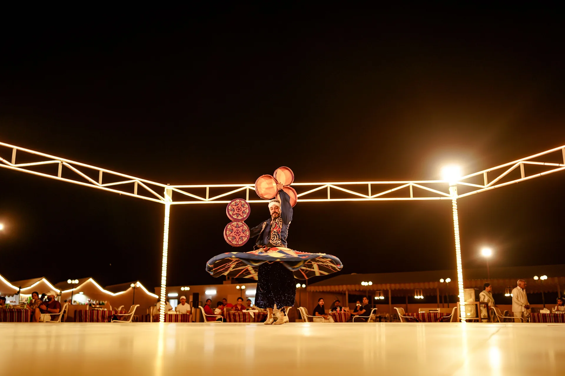 Une danseuse de Tanoura tourne sous une arche d'acier illuminée sur une scène en plein air lors d'un dîner-spectacle dans le désert, avec des tables de restaurant et des pavillons sous tente visibles en arrière-plan (photographie représentative d'une représentation de Tanoura dans le désert des Émirats arabes unis, et non de la scène propre à Al Hadheerah)